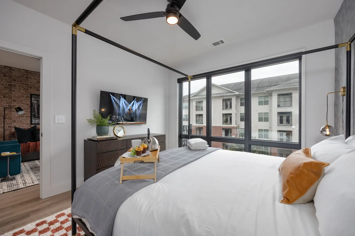A bright, modern bedroom featuring a bed with crisp white linens and decorative pillows, a television mounted on the wall, a breakfast tray on the bed, and large windows flooding the space with natural light. The view outside hints at the city beyond, while the clean lines and subtle accents create a peaceful, upscale atmosphere.