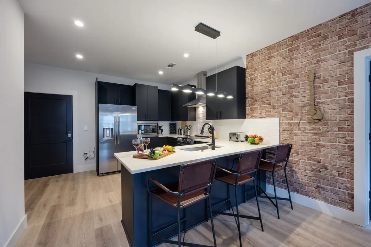 A welcoming kitchen interior showcasing a kitchen island with bar stools, exposed brick wall, and countertop accessories. The space is styled for both casual dining and entertaining, with a layout that promotes conversation and community.