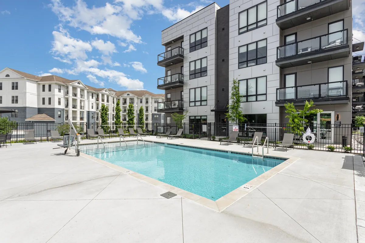 An inviting exterior shot featuring the sparkling swimming pool, surrounded by residential buildings, lush landscaping, and patio furniture. The relaxed atmosphere is ideal for sunbathing, swimming, or chatting with neighbors.